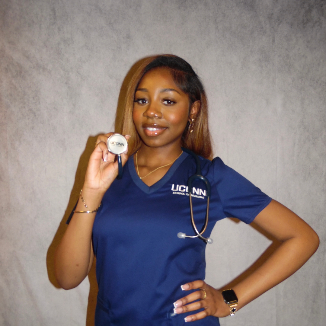 A woman in navy blue scrubs holding a stethoscope and smiling, standing against a plain background.