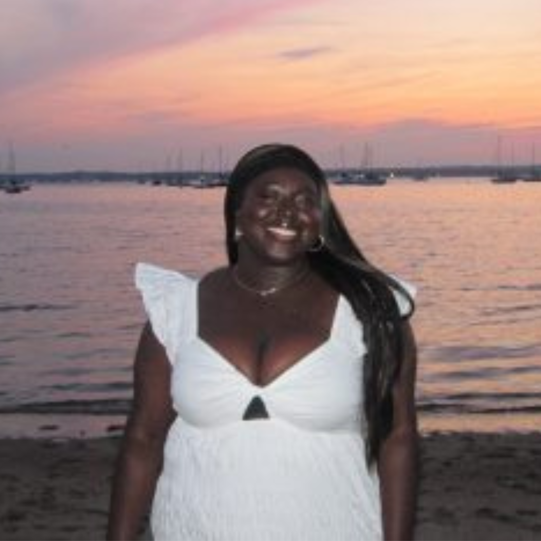 Student is smiling at the camera. She is wearing a white dress, has long black hair, and is posed in front of a pink ocean sunset.