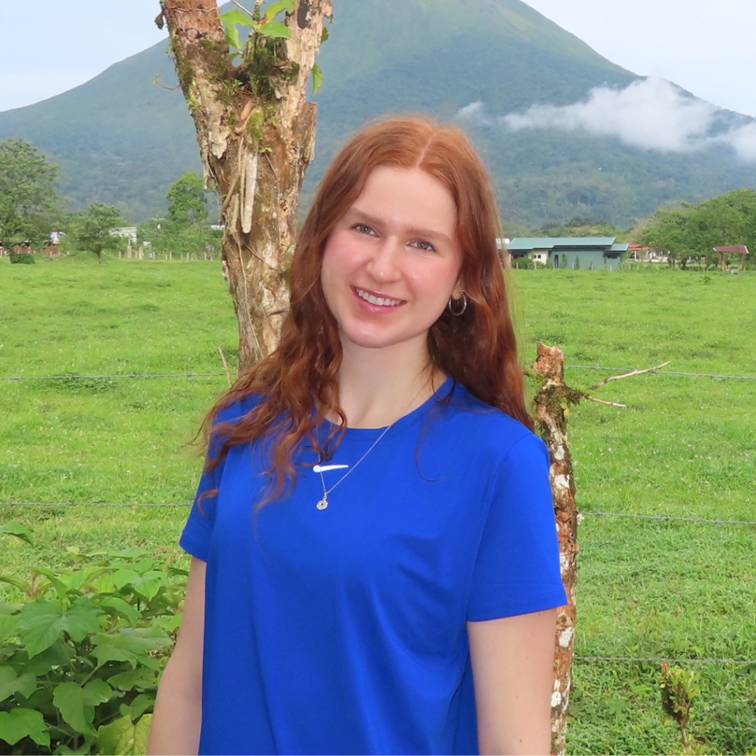 Headshot of a UConn student. She has medium length red hair, is wearing a blue t-shirt, and is posed in front of a grassy mountain.
