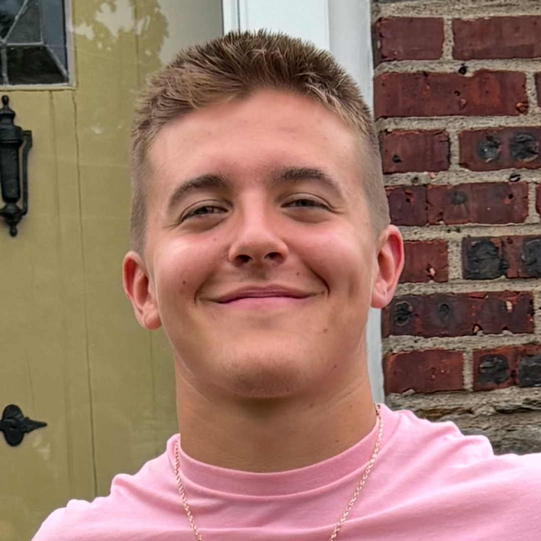 A young man smiling outdoors, wearing a pink shirt and standing in front of a brick wall and door. 