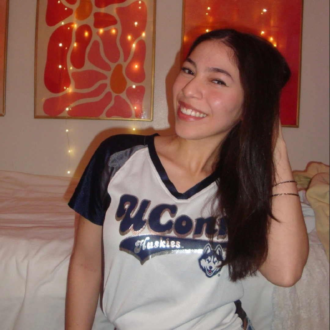 Photo of UConn student. She has medium black hair, is wearing a UConn t-shirt, and is posed with her hand in her hair in her dorm.