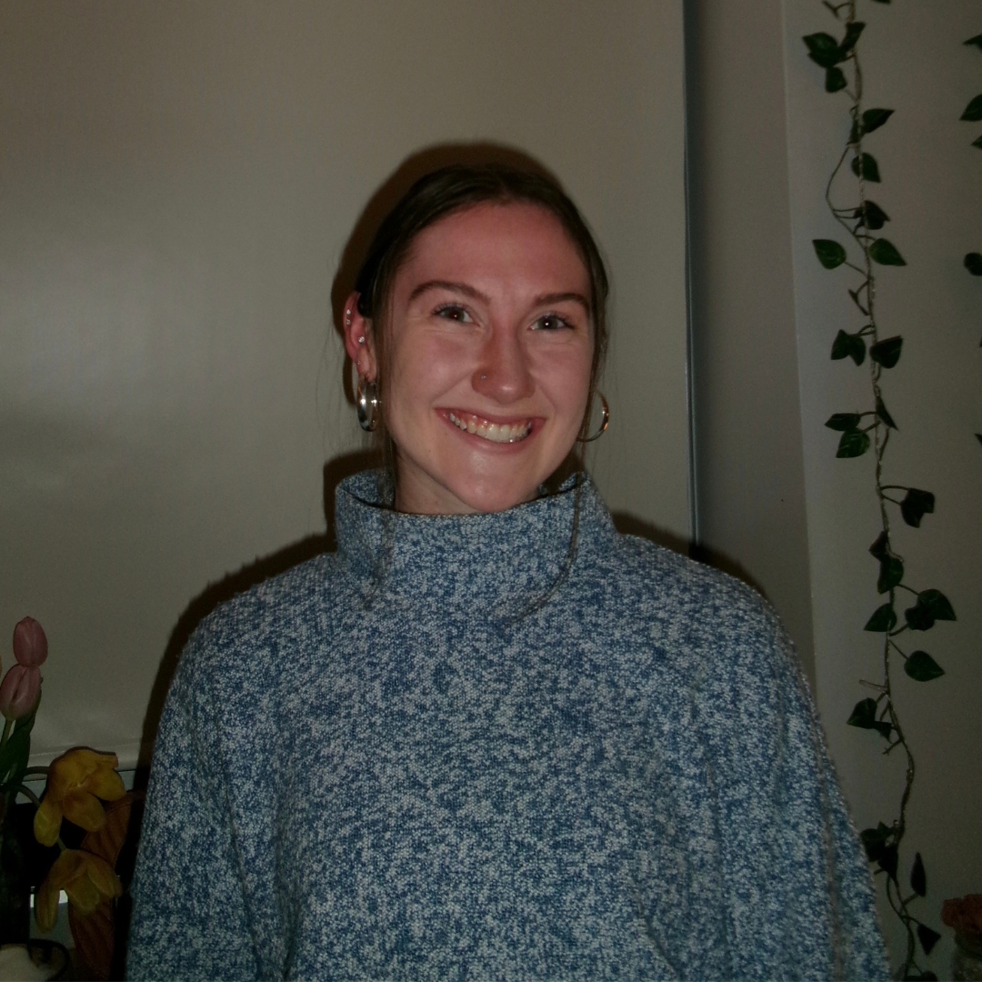 A woman smiling indoors, wearing a blue sweater with plants and flowers in the background.