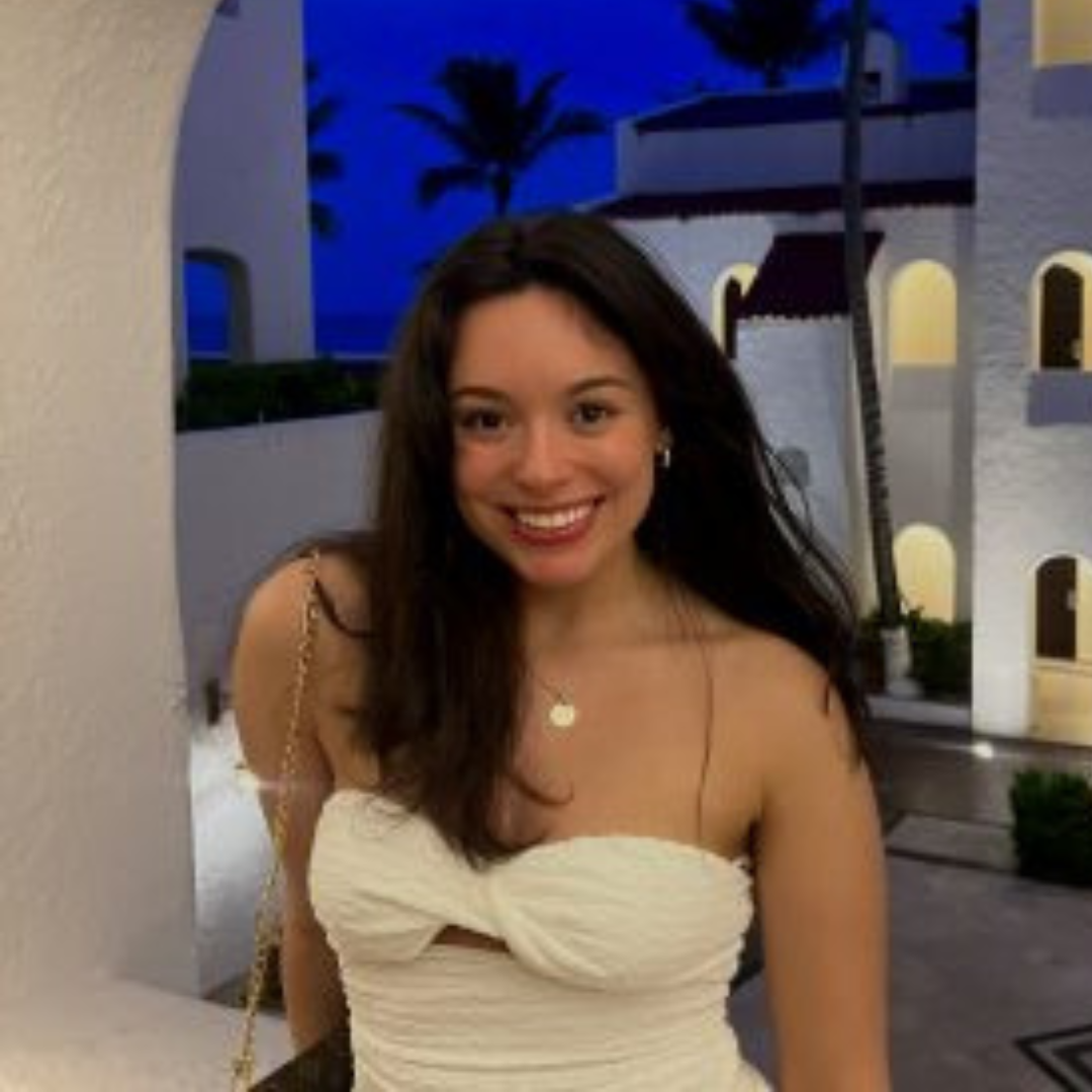 Student is smiling at the camera. She is wearing a white dress and is posed in front of white buildings and palm trees, and a vivid blue sky.
