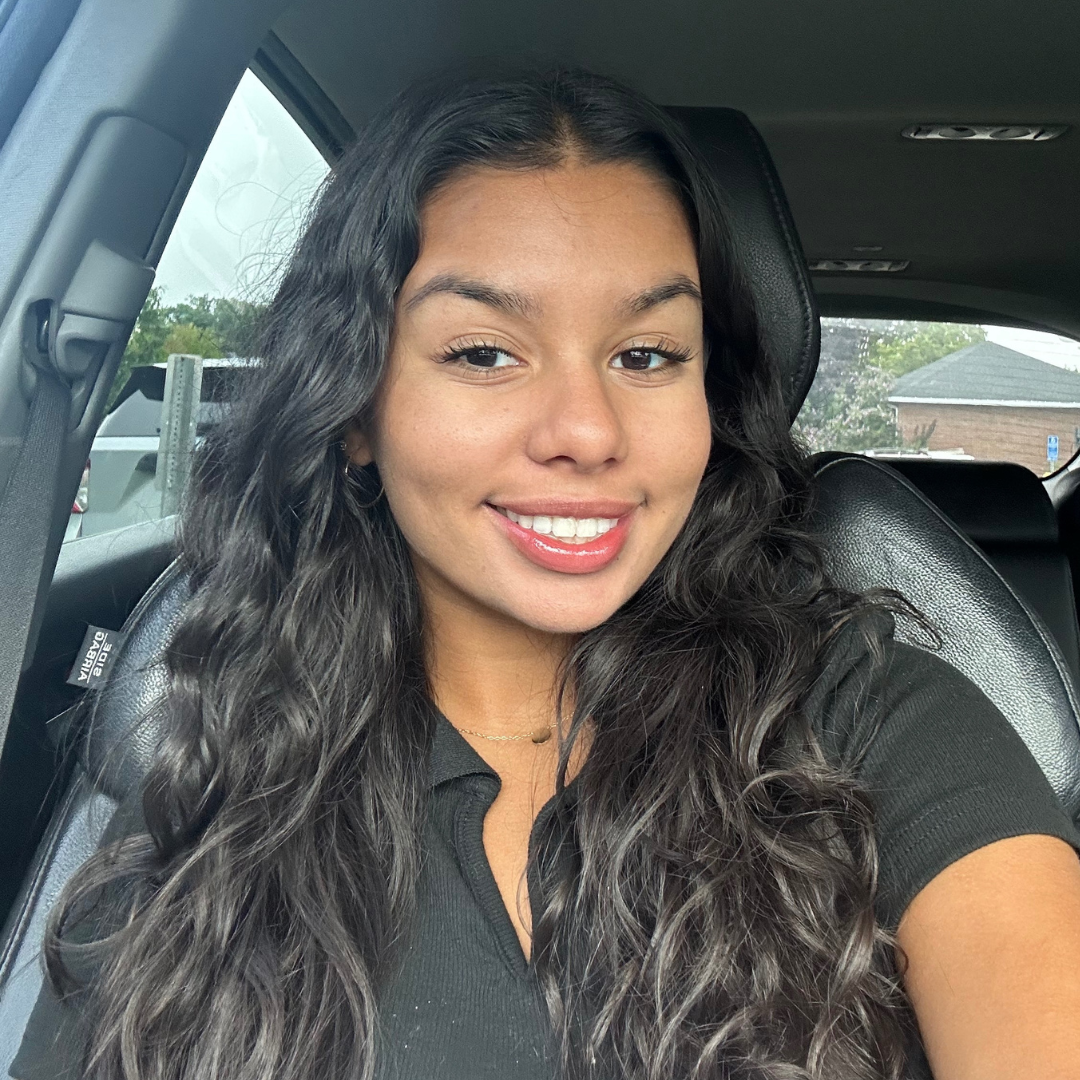 Headshot of UConn student. She has long black hair, is smiling at the camera, is wearing a black shirt, and is posed in the car.