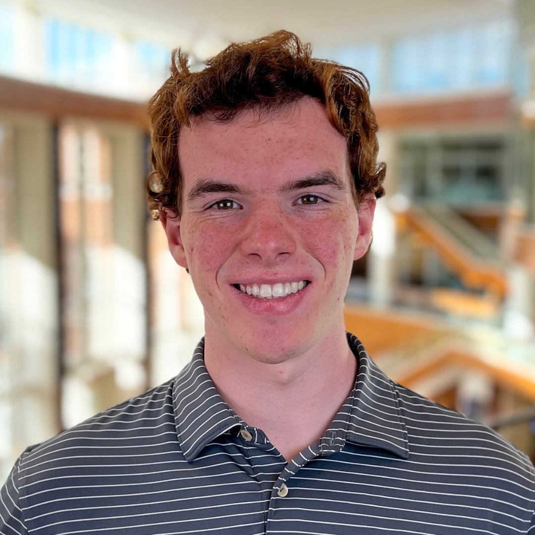 A young man smiling indoors, wearing a striped polo shirt, with a bright and modern building interior in the background,