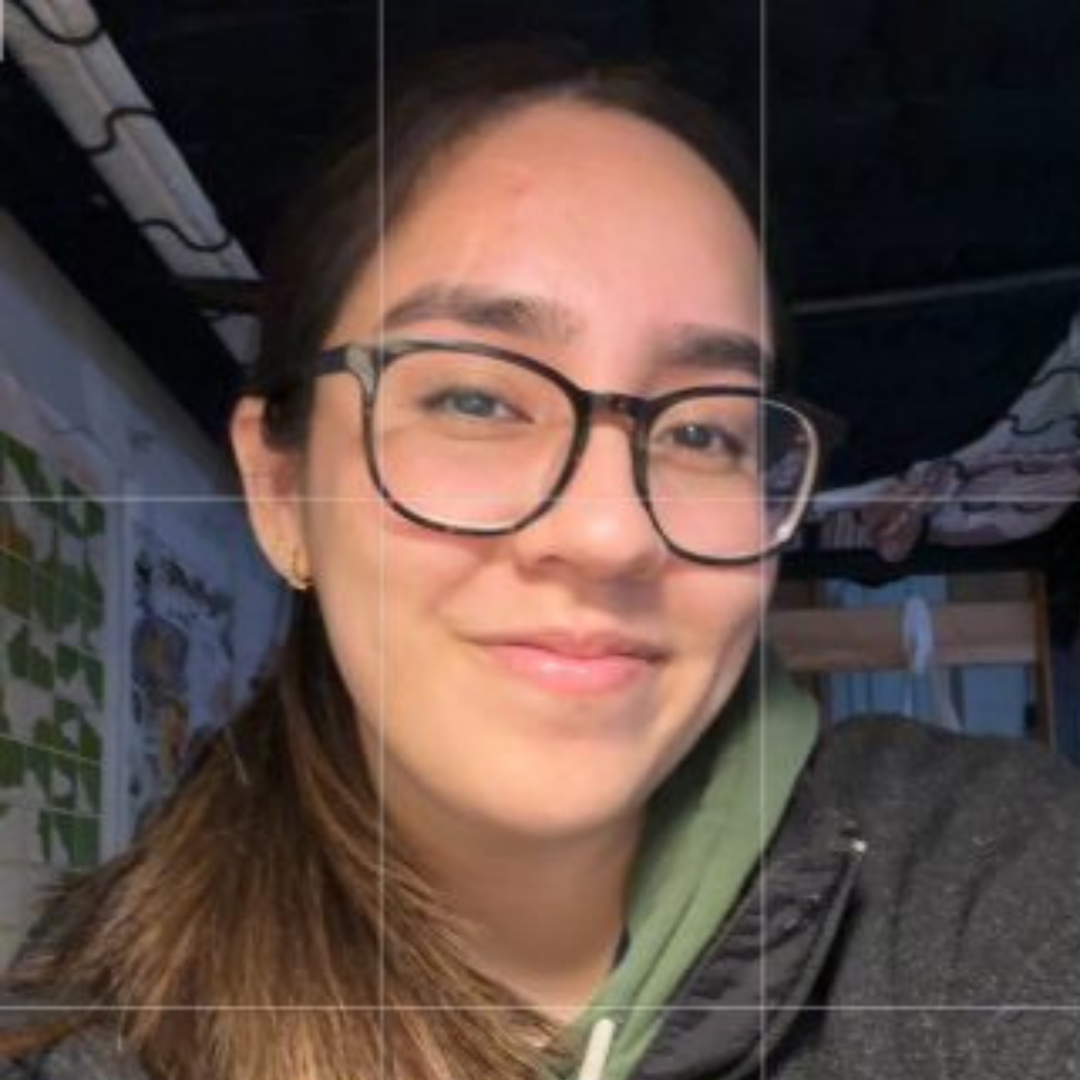 Student is smiling at the camera. She is wearing a green hoodie under a black jacket, black glasses, and her brown hair in a ponytail. She is posed in her dorm room.