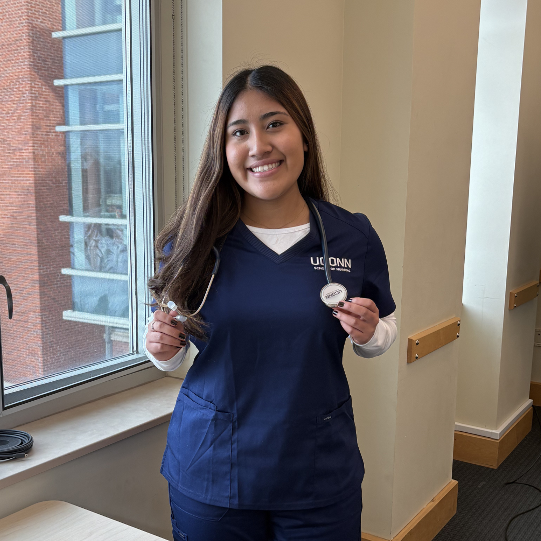 A woman in navy blue scrubs smiling and holding a stethoscope, standing by a window in a bright indoor setting.