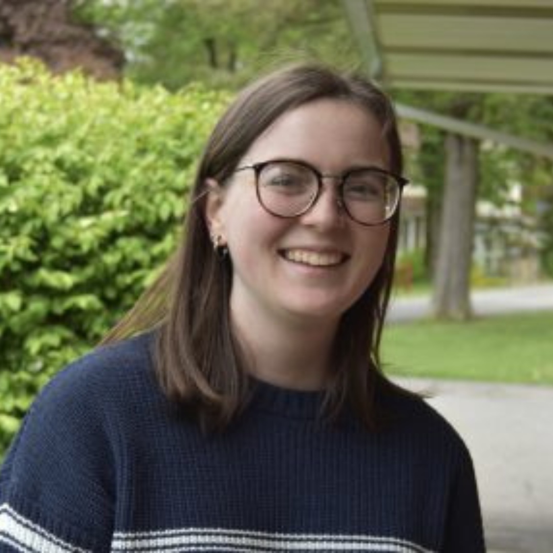 Student is smiling at the camera. She is wearing a blue sweater with a white stripe, black glasses, and has short brown hair. She is posed outside in front of a green landscape.