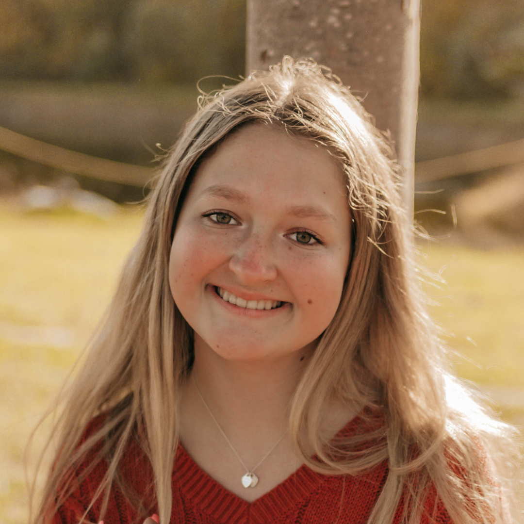Headshot of UConn student. She has long blonde hair, is wearing a red blouse, is smiling at the camera, and is posed in front of a field.