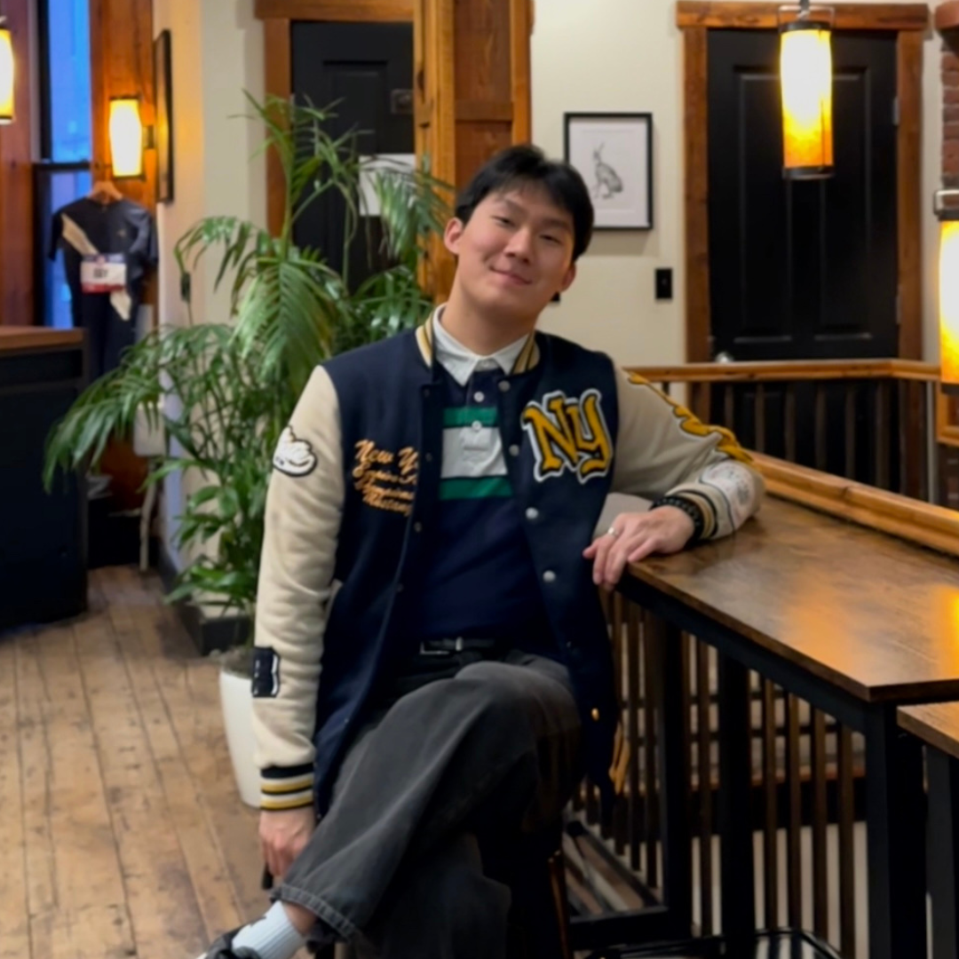 A man smiling and leaning on a wooden railing indoors, wearing a varsity jacket and sitting in a warmly lit space with plants and artwork in the background.