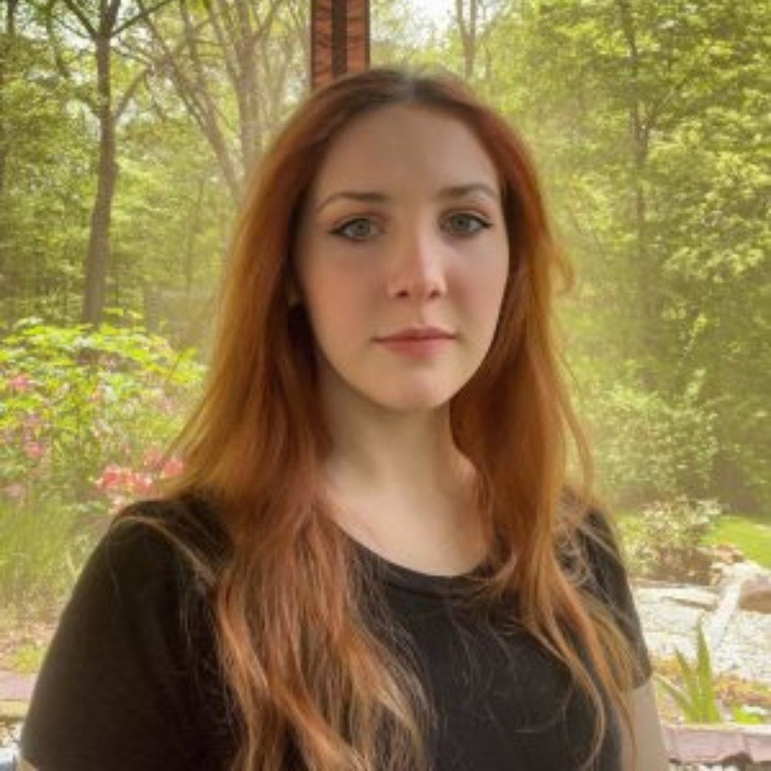 Student is smiling at the camera. She is wearing a black long-sleeved shirt, and has long red hair. She is posed in front of a window with a forest landscape.