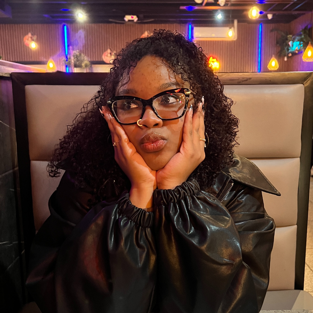 A woman with curly hair and glasses resting her face in her hands, seated in a booth at a warmly lit restaurant. 