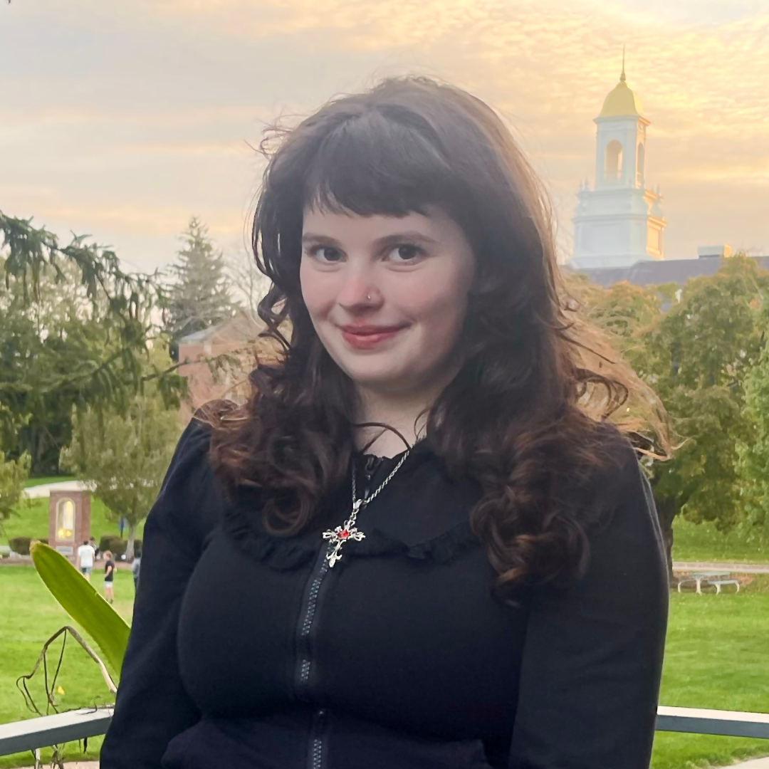 Photo of UConn student. She has short curly black hair, is wearing a black blouse, is posed in front of Wilbur Cross, and is smiling at the camera.