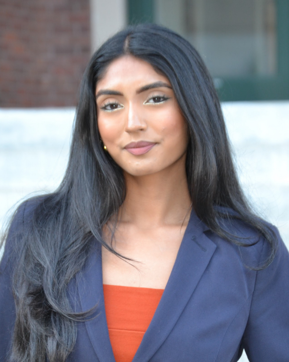 Headshot of student worker. She is wearing a navy blue blazer, bright orange blouse, and has black hair.