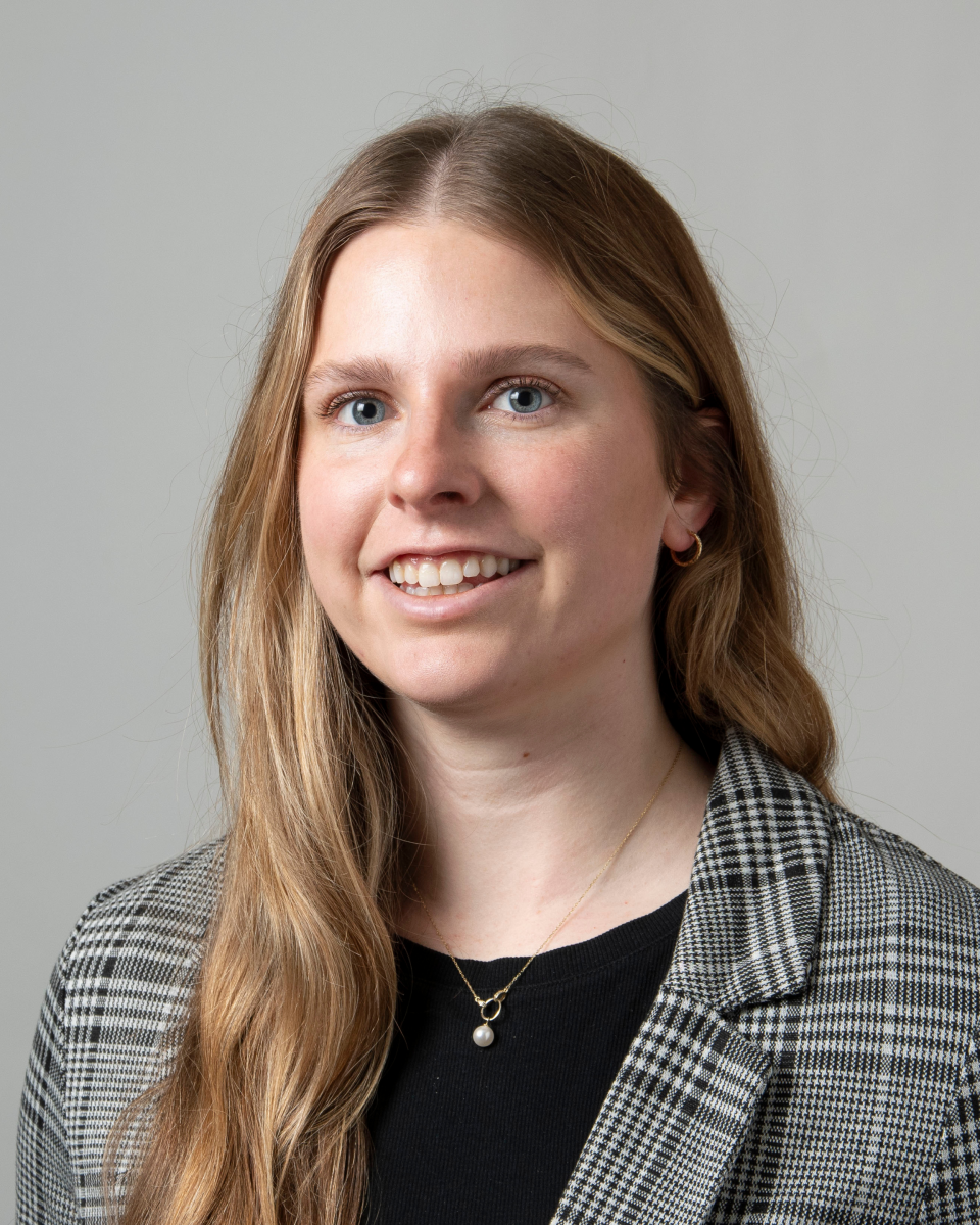 Photo of staff member smiling at the camera. She has long blonde hair, and is wearing a black blouse, with a checkered blazer.