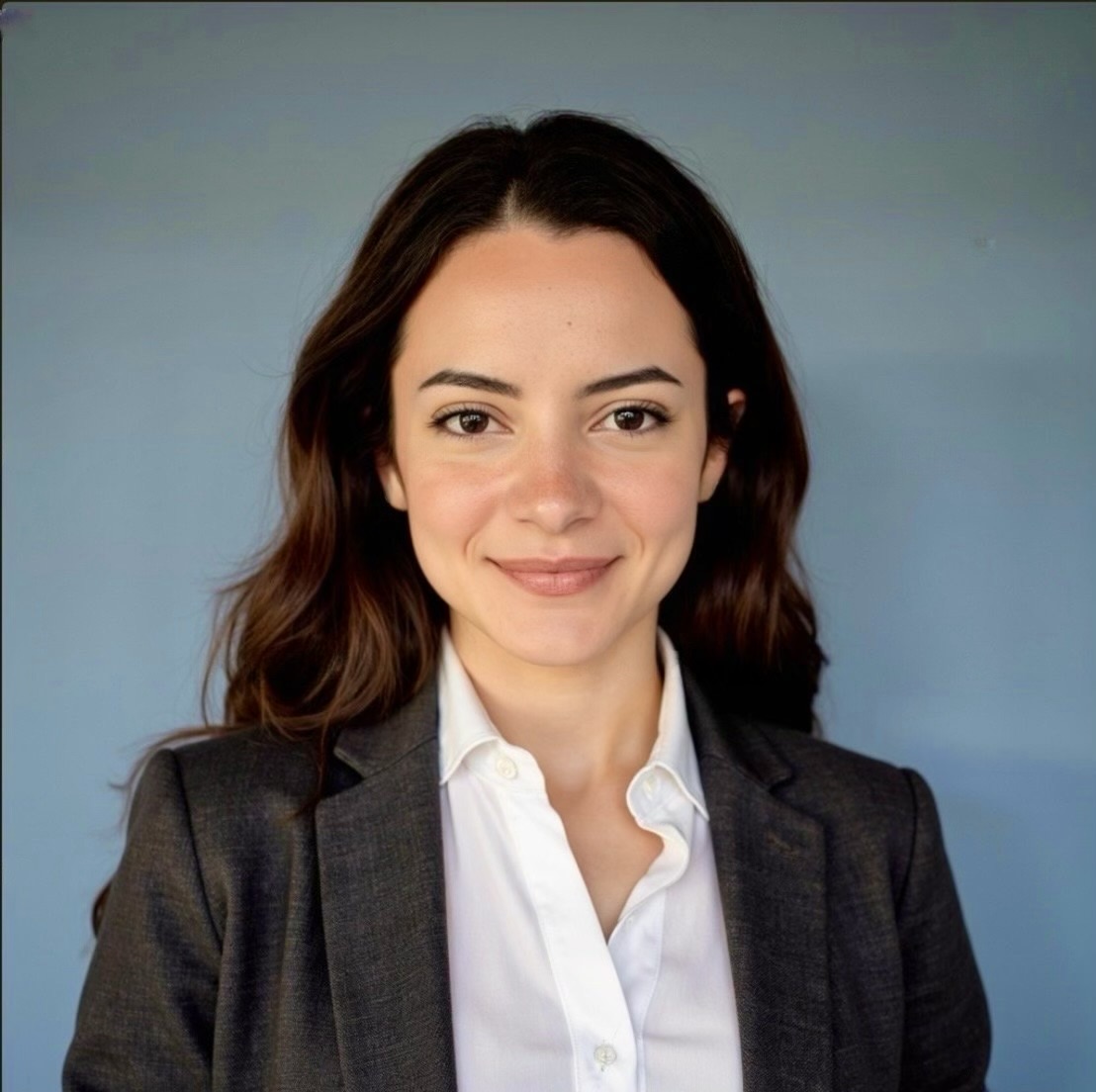 Photo of a young white woman with medium length brown wavy hair. She is wearing a gray blazer with a white shirt underneath