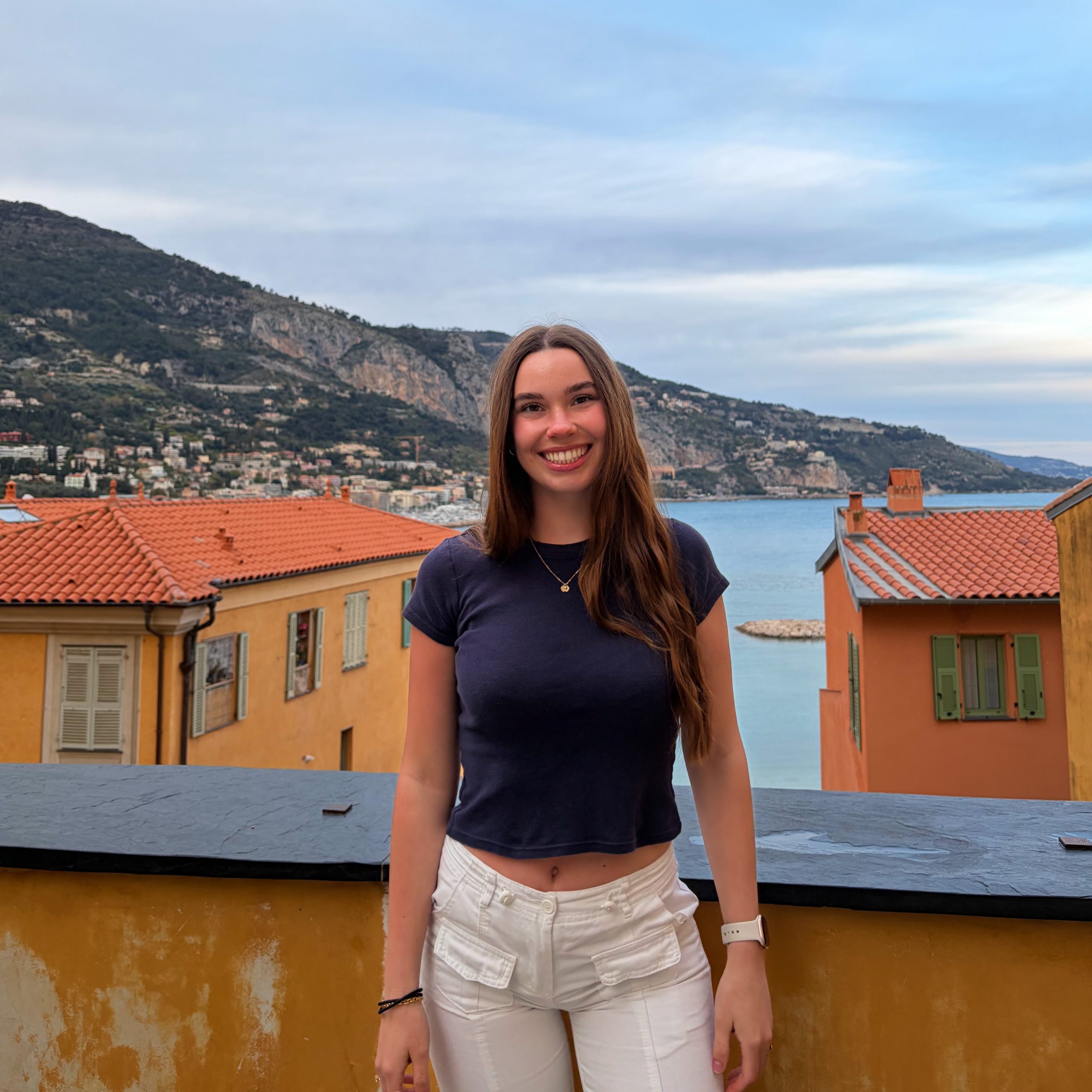 Photo of a young woman smiling towards camera with long brown hair wearing a blue t-shirt and white pants. She is standing in front of a barrier with a town and sea in the background.
