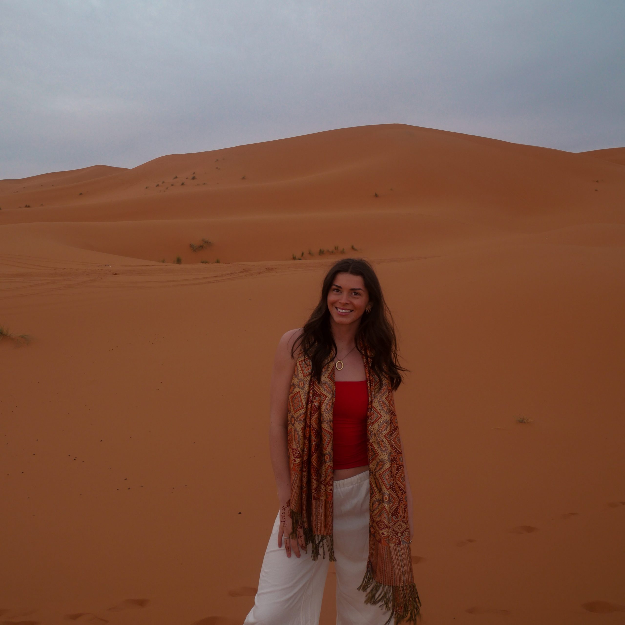Photo of a woman with medium length brown hair wearing a red shirt and white pants with a scarf in the Sahara Desert.