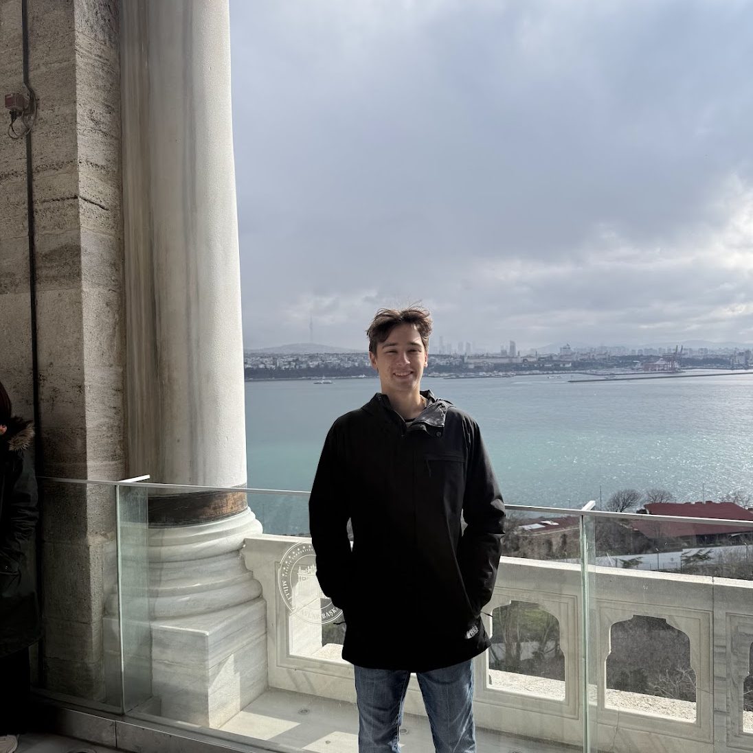 Photo of a young man wearing a black wind breaker and blue jeans in front of a glass barrier with the sea and a city in the background.