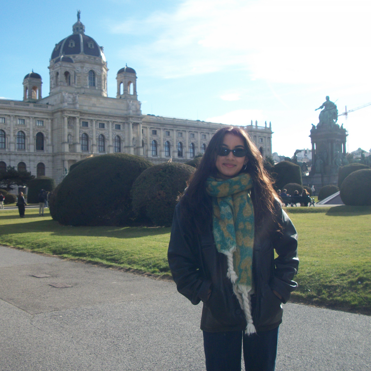 Photo of a young woman with long brown hair wearing sunglasses and a green scarf smiling towards camera. There is a building with a dome and statues in the background, along with green patches.