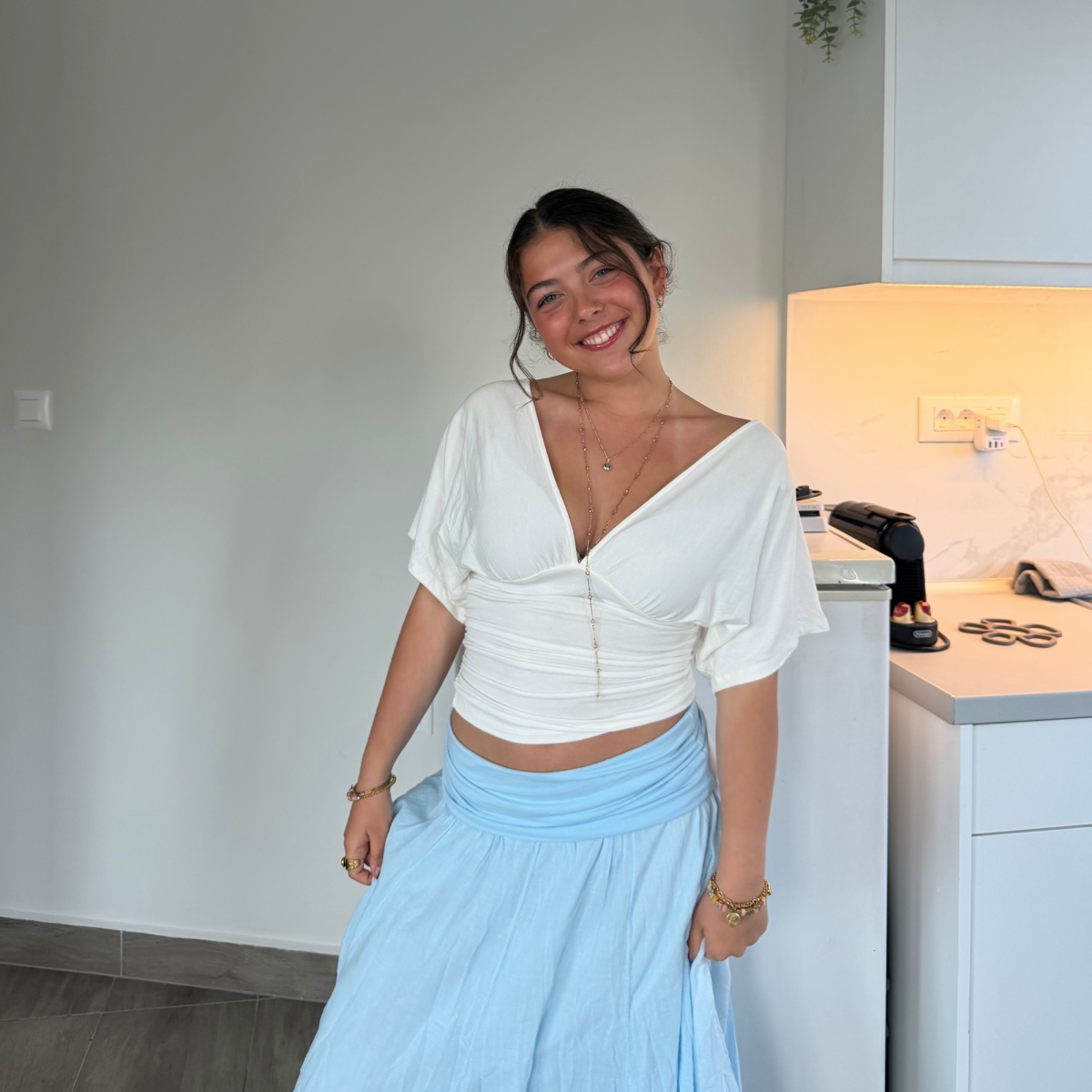 Photo of a young woman smiling towards camera with dark hair tied back. She is wearing a white blouse with a light blue loose skirt in front of a white wall.