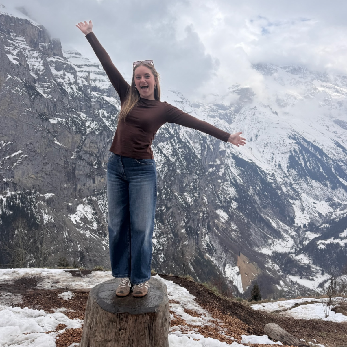 Photo of a young woman with medium length blonde hair smiling with her arms stretched out wearing a brown long sleeve shirt and dark blue jeans in front of a snowy mountain top.