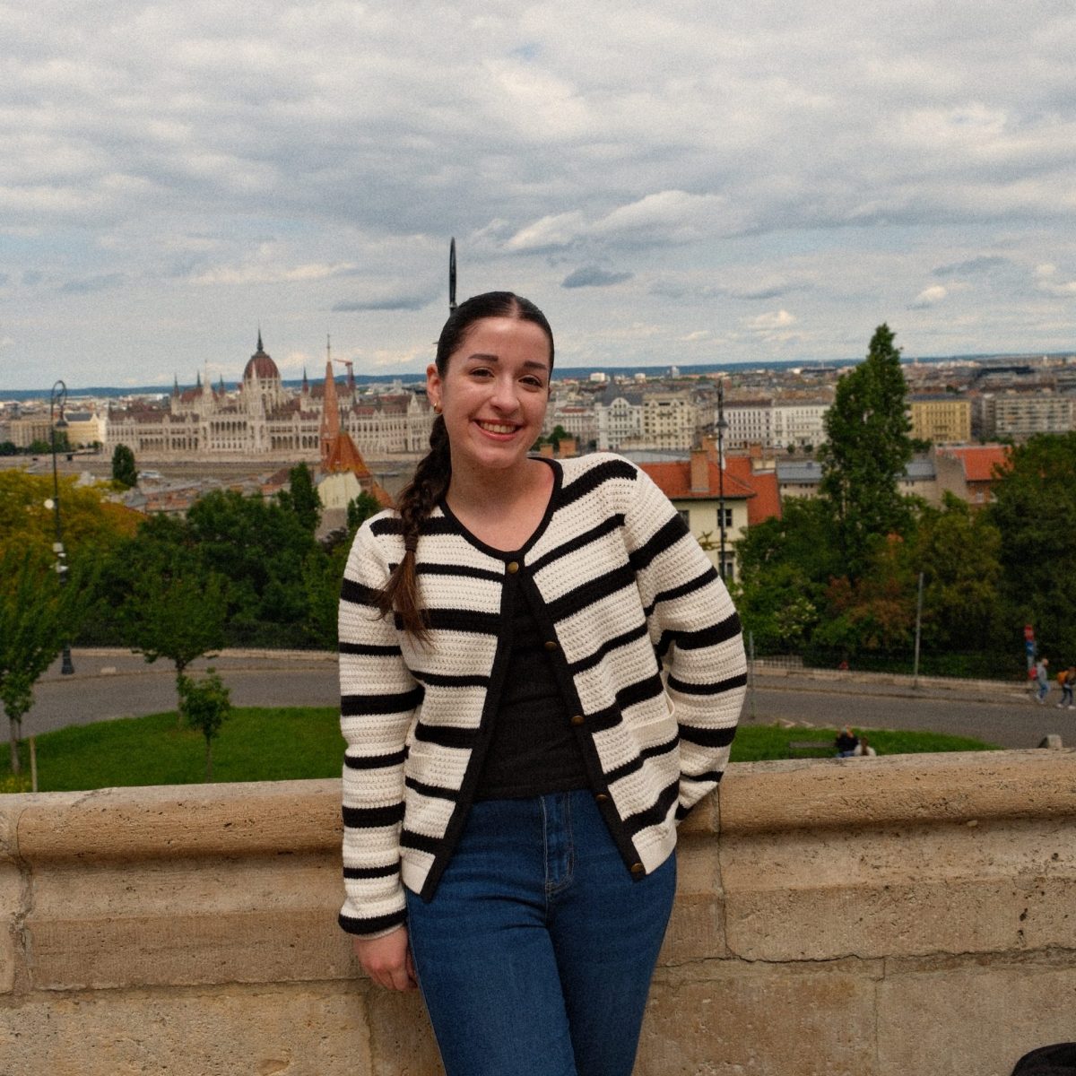 Photo of a female student with braided brown hair wearing a white and black striped cardigan in front of a city.