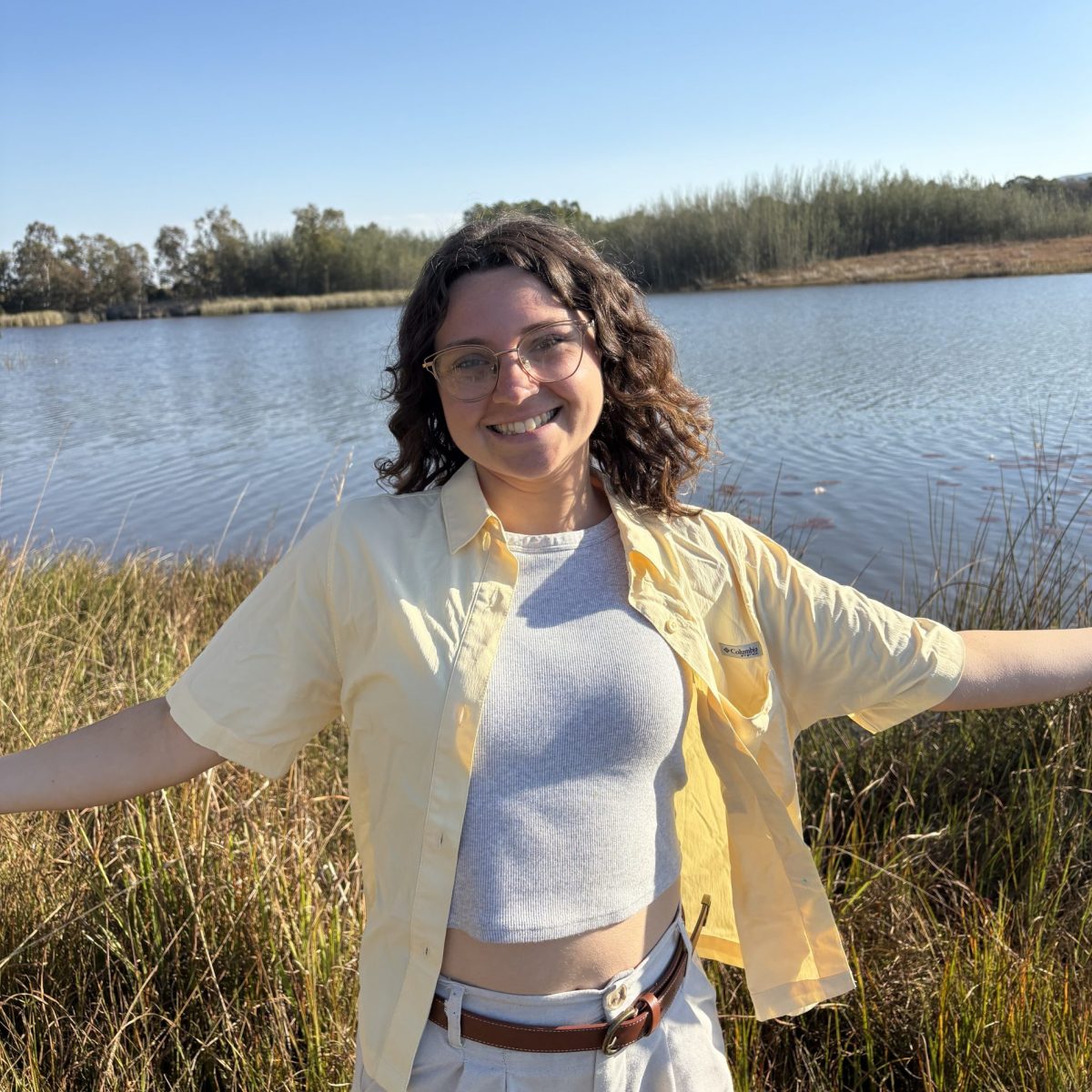 Photo of a female student wearing a yellow button up shirt with a white top underneath smiling with her hands out in front of a pond.
