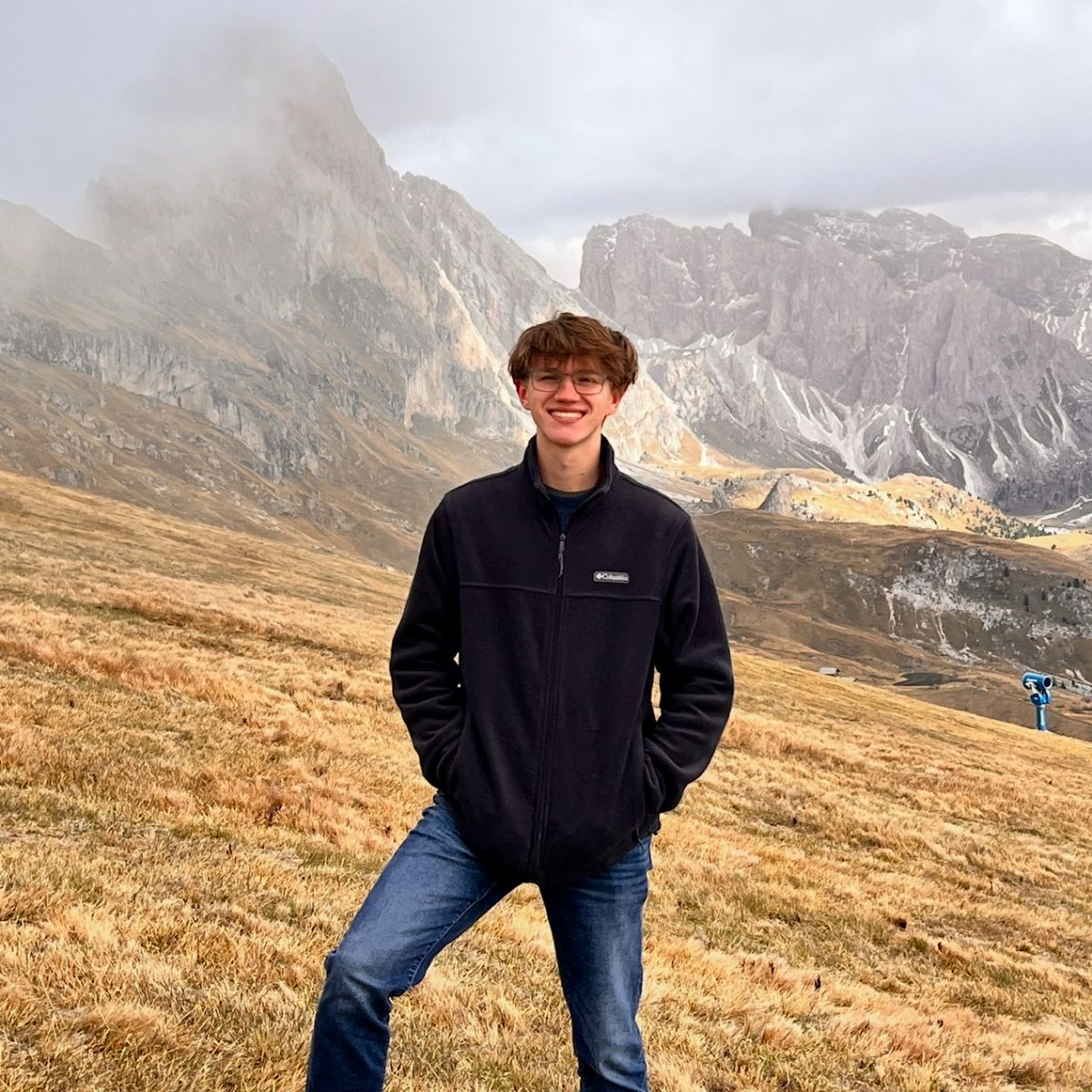 Photo of a male student wearing a black zip up jacket with mountains in the background.