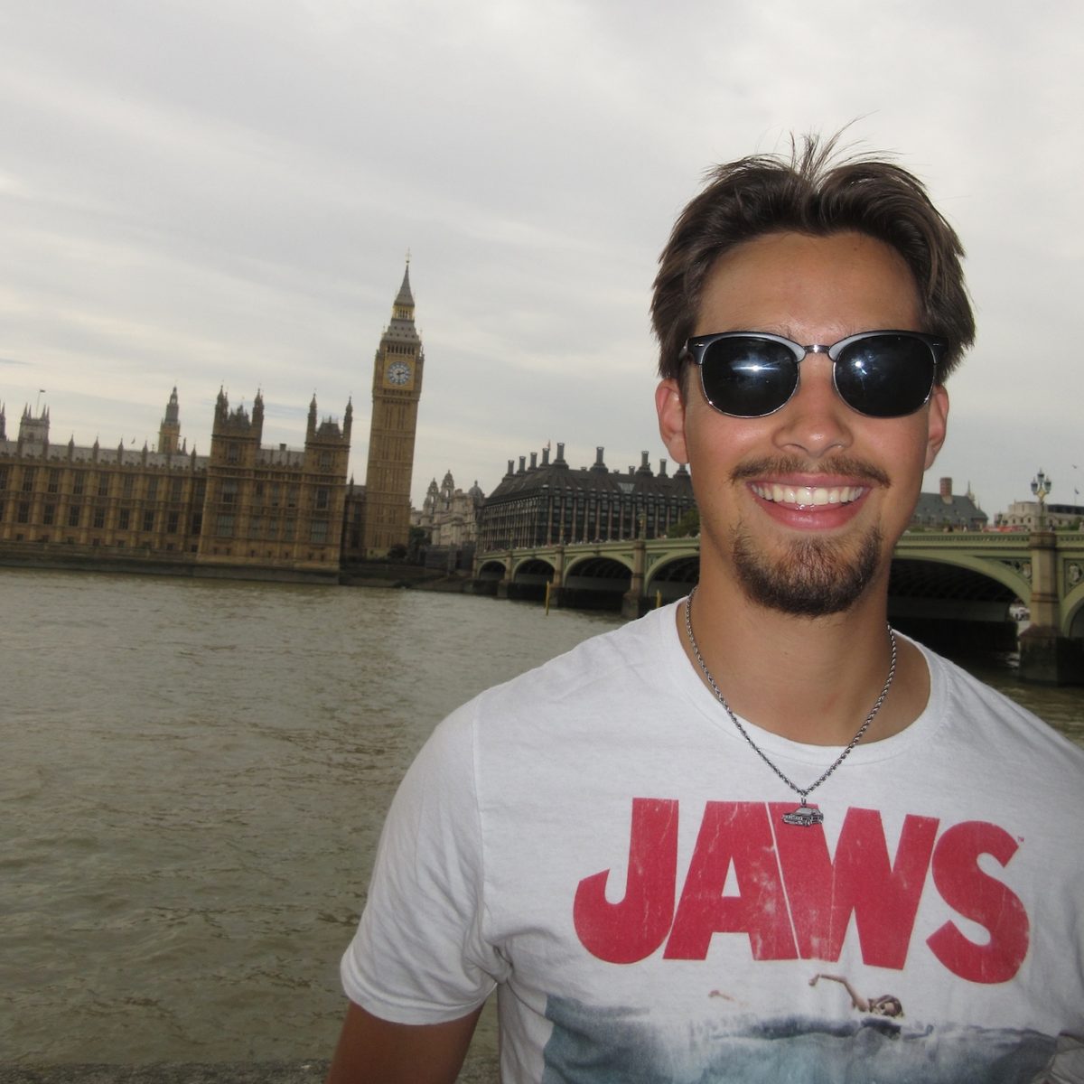 Photo of a male student wearing sunglasses with a white t-shirt that reads JAWS with the London skyline in the background.