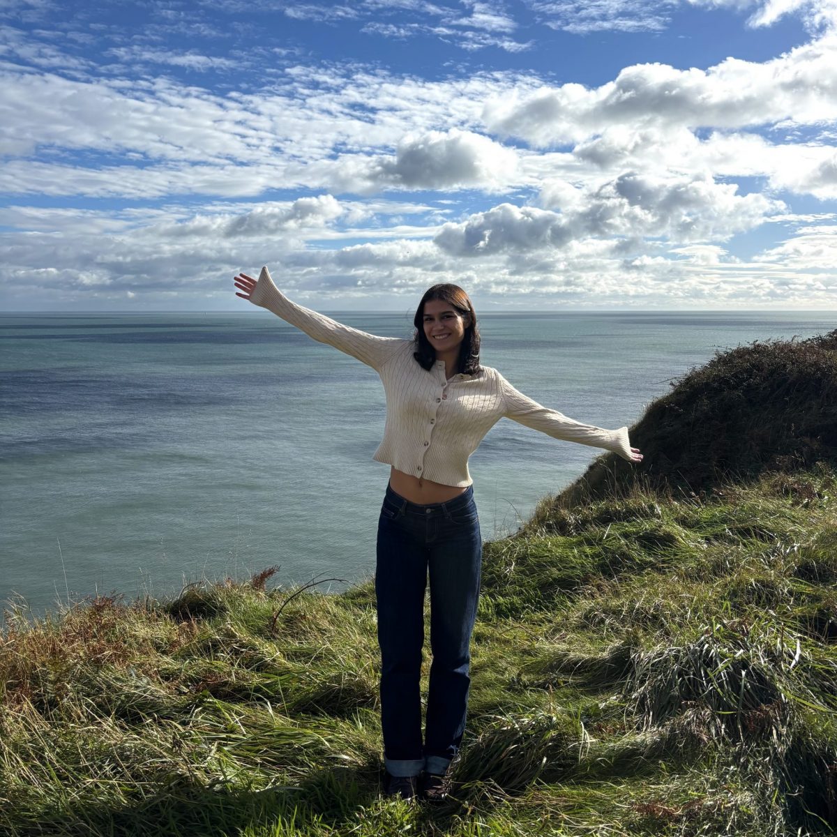 Photo of a female student wearing a white cardigan and jeans with her hands out on a grassy patch in front of the sea.